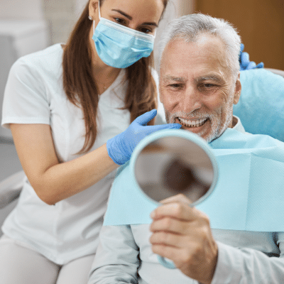 Implant restorations in asheboro nc: restoring the function and beauty of your smile 6 Older man looking at his teeth in a small mirror, representing implant restorations in asheboro, nc